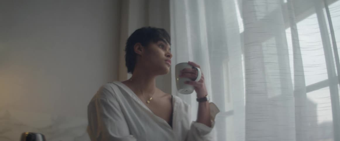 A young woman holding a coffee cup walks across a home kitchen to a window and gazes out contemplatively