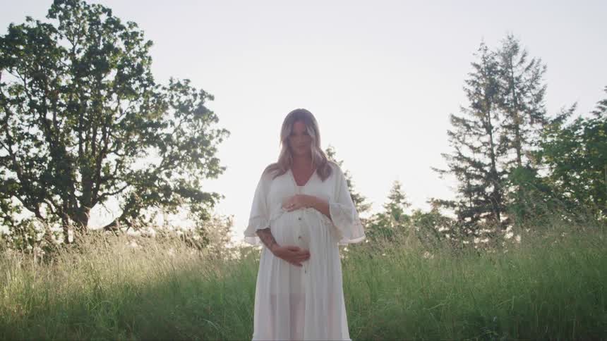 A woman has her hands placed around her pregnant stomach while standing in tall grass
