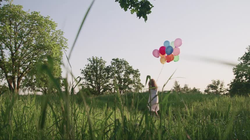 Young girl with colorful hellium balloons walking on the green grass in a field at evening