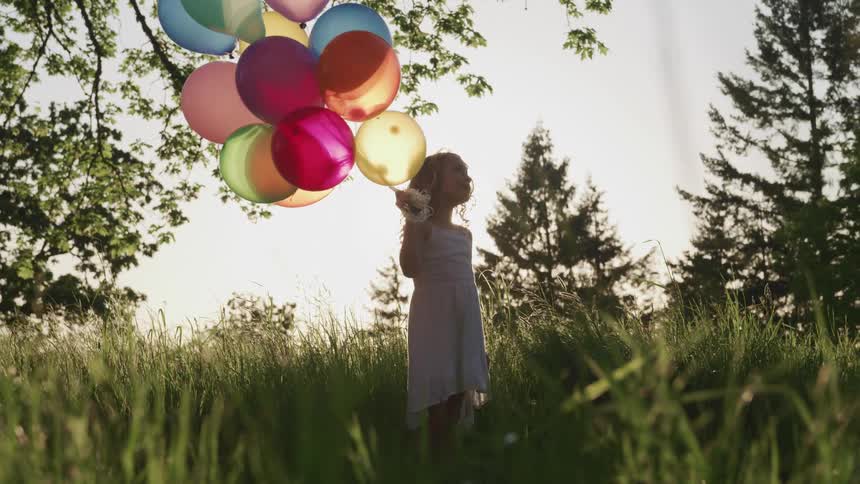 Young girl wearing a dress holding a bunch of colored balloons on a sunny day with trees all around