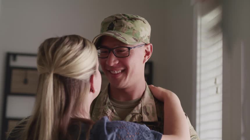 A man smiling wearing army clothing hugs his wife at the entrance of a home
