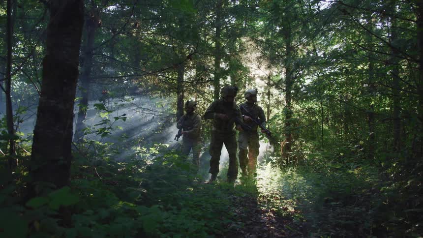 Three military soldiers wearing helmets move through the forest with their guns in hand as the sun shines brightly behind them