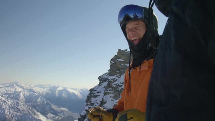 Two men wearing ski gear stand on top of a mountain summit smiling at daytime
