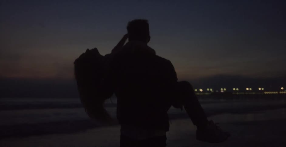 Man carrying a woman in his arms on the sand on a beach near an amusement park at night