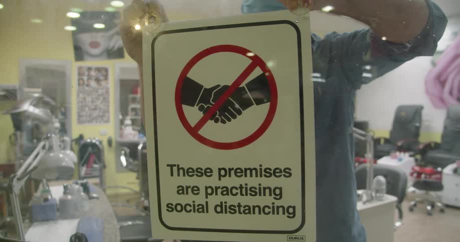 White man putting up a sign about social distancing at the glass door of his hair salon