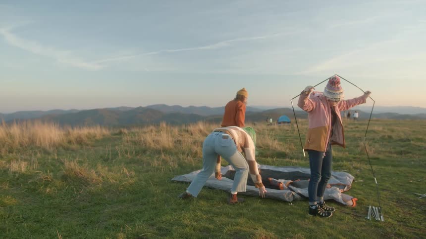 Group of female friends setting up a tent for camping in the mountain at sunset