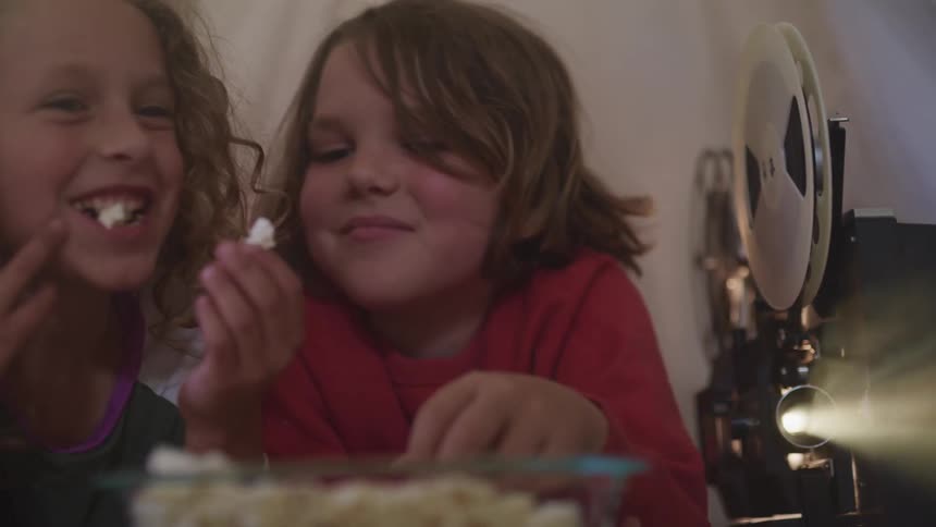 Two kids smiling laughing and eating popcorn next to a film projector