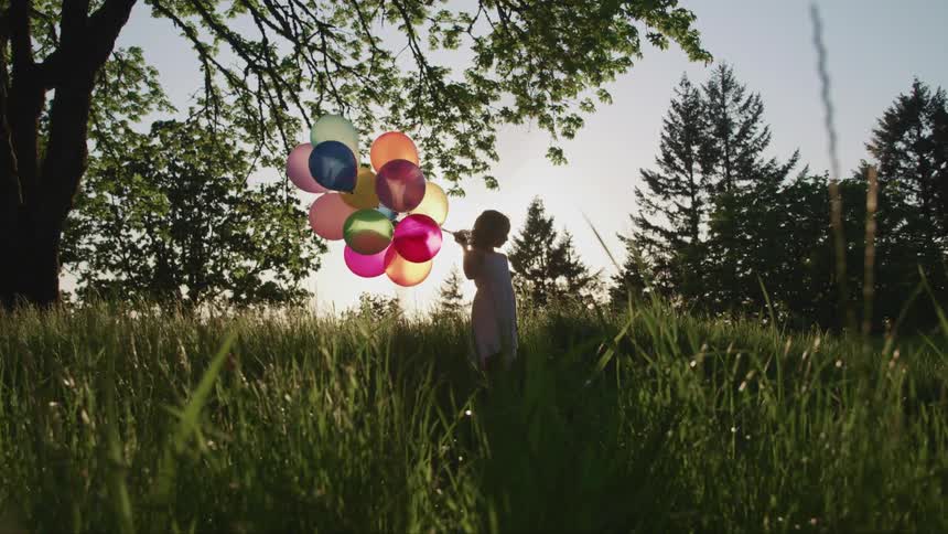 Young girl wearing dress holding a bunch of colored balloons while standing in tall grass with many trees behind her