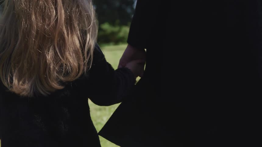 An adult holding a childs hand walking in a graveyard both wearing black
