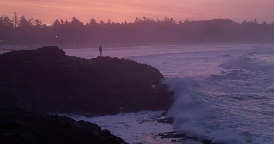 Person standing on rocky shoreline as waves roll in splashing with trees on the horizon and a red sky