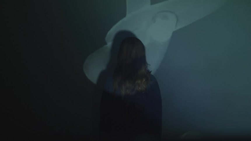 Woman stands with her face towards a wall while shadows and lights with different symbols are casted around her