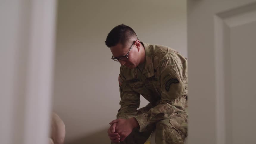 Man in a military uniform and wearing glasses sits on the edge of a bed