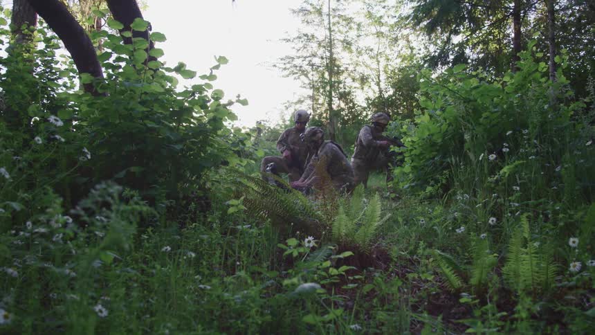 Two military soldiers crouched day working on a computer device while a third soldier taking watch with a gun