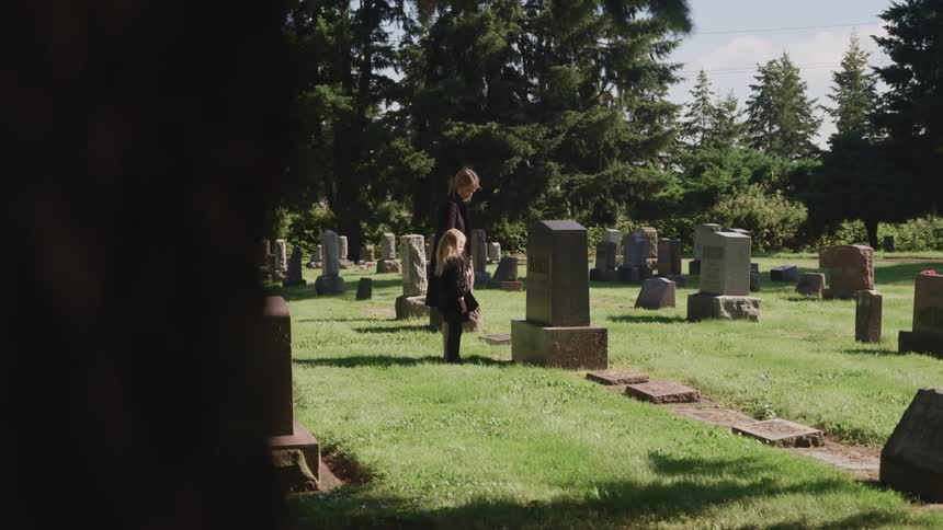 A woman and a girl standing in a cemetery with trees the sky and clouds in the background