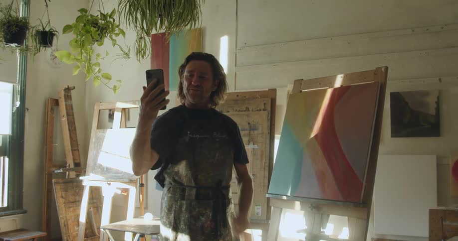 Man standing in front of an easel with a painting in an art studio on a video call on a smart phone at daytime