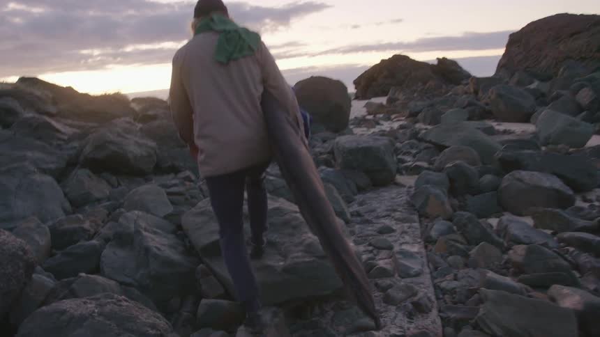 Man holding a bag with a surfboard climbing up piles of rocks walking towards a beach at sunset