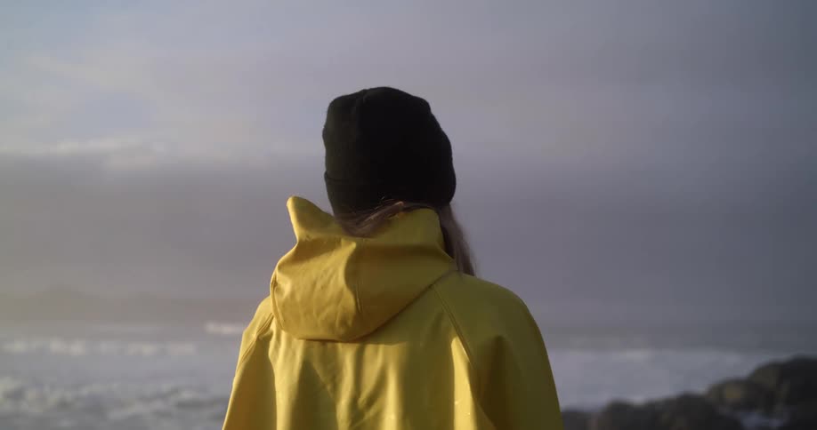 A woman stands at the shoreline looking at the waves while wearing a yellow raincoat and knit hat