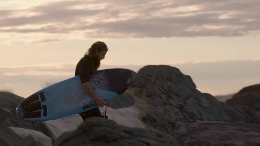 Man holding a surfboard while walking down the rocks at the beach