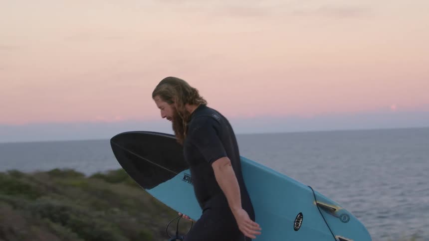 Man walks along a beach carrying a blue surfboard