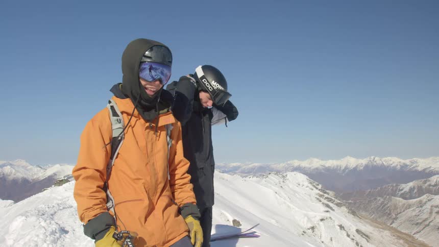 Two men preparing their ski gear at the top of a snowy mountain summit at daytime