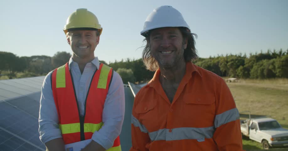 Two construction workers wearing hard hats smiling at the camera from the rooftop of a building