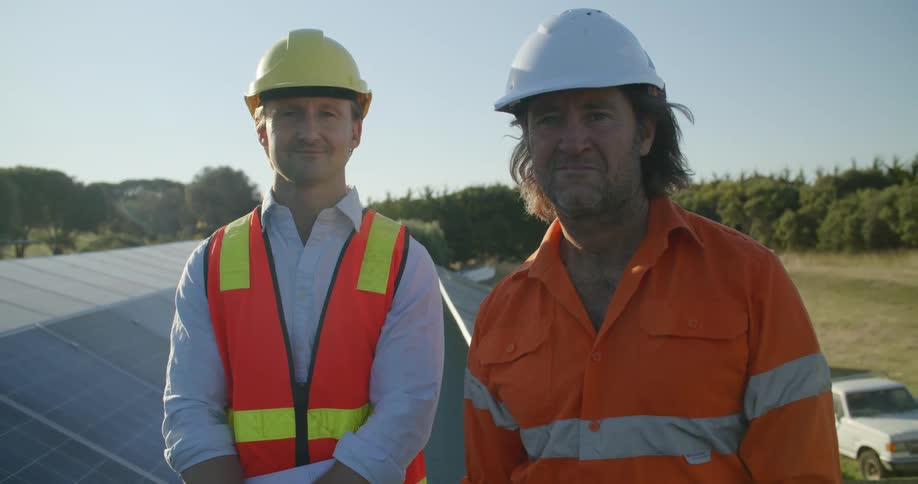 Two men dressed in safety gear on top of a roof smile at the camera