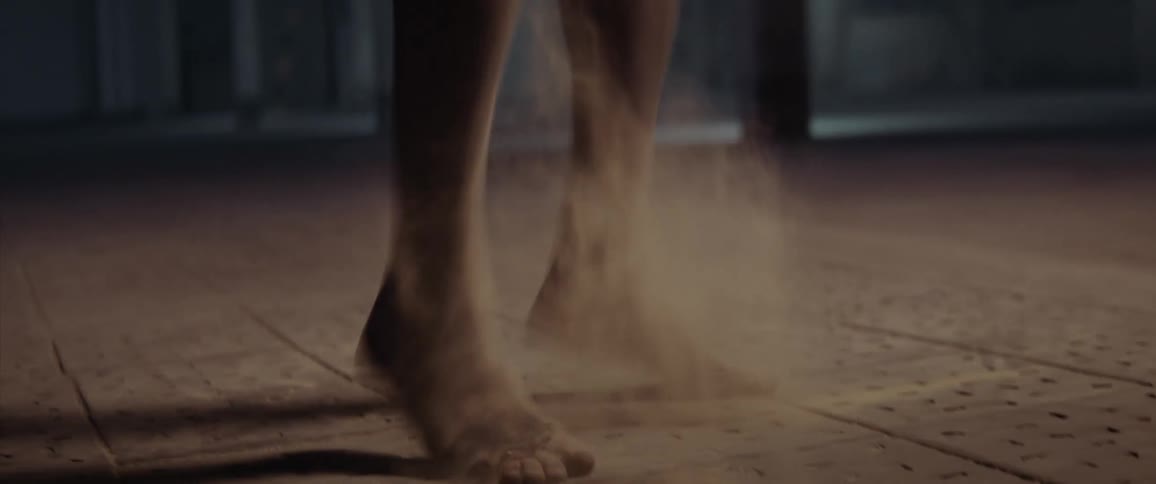 A woman with dust over her feets jumps and falls down on the floor of a dimly lit room