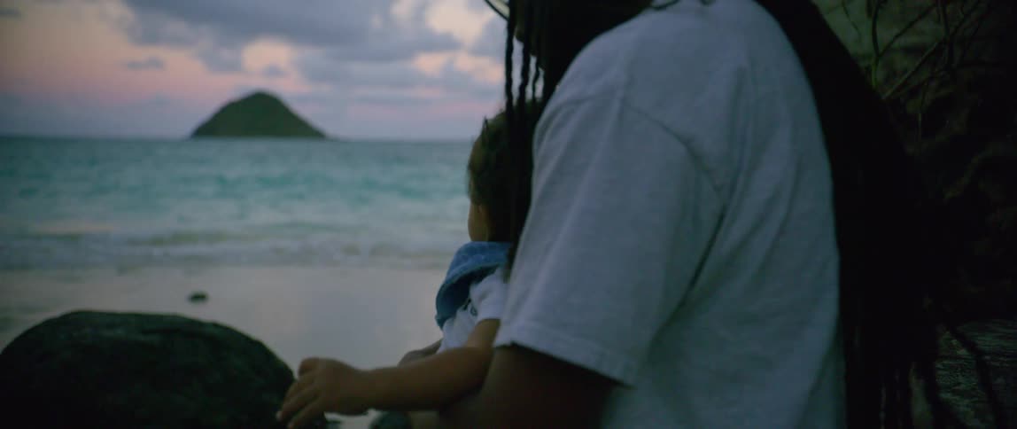A man with a hat and glasses sitting and holding a baby on the beach with the ocean sky and clouds in the background