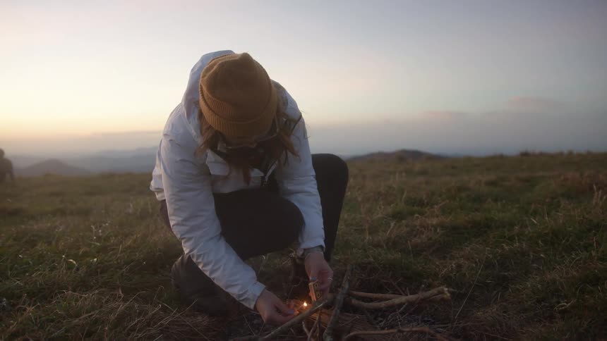 Woman crouches down in a field and strikes a match to start a fire with some sticks