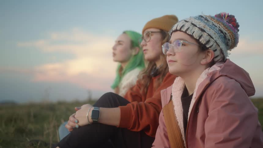 A young woman gestures out of frame and speaks to her sisters as they sit together in the grass wearing cold weather clothing during the day