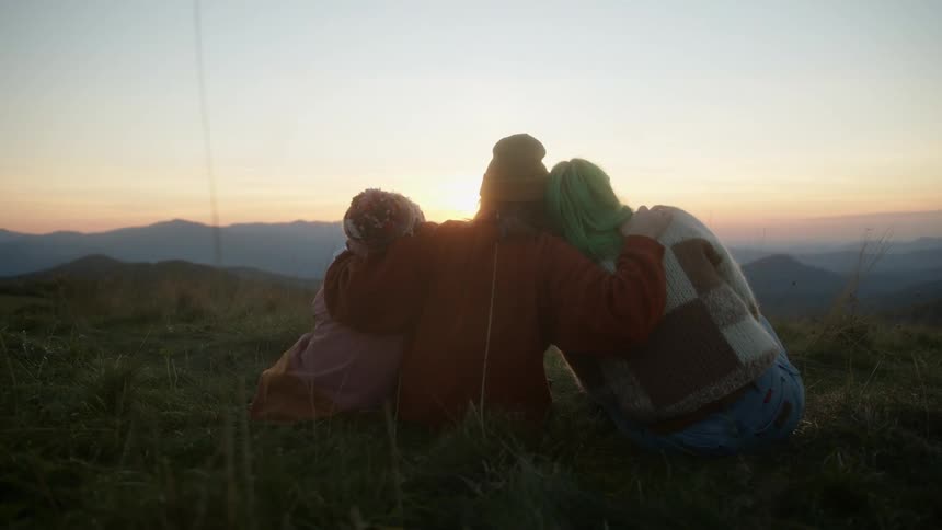 A group of female friends sitting atop a mountain hugging while looking at the sunset
