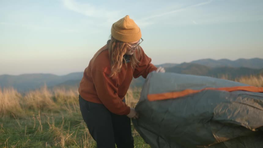 Young blonde woman assembling a tent camping on the mountain at sunset