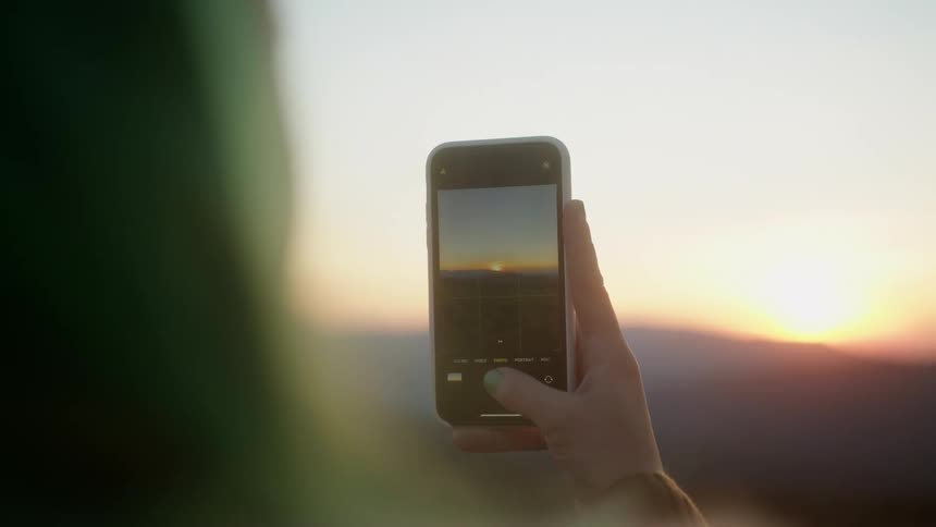 Woman takes a photo with a smartphone of the sun setting behind the mountains