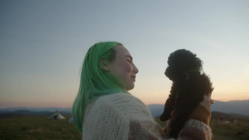 A young woman holding a puppy up in the air then kissing and hugging the puppy with grasslands and mountains along the horizon