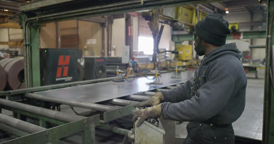 A man in a factory watches a sheet of metal on a conveyor belt