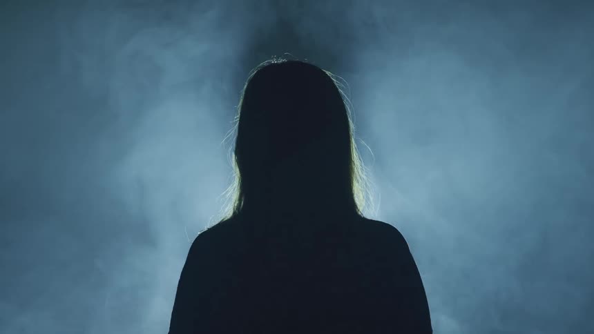 Silhouette of a woman standing in front of a strobing white light in a room filled with smoke