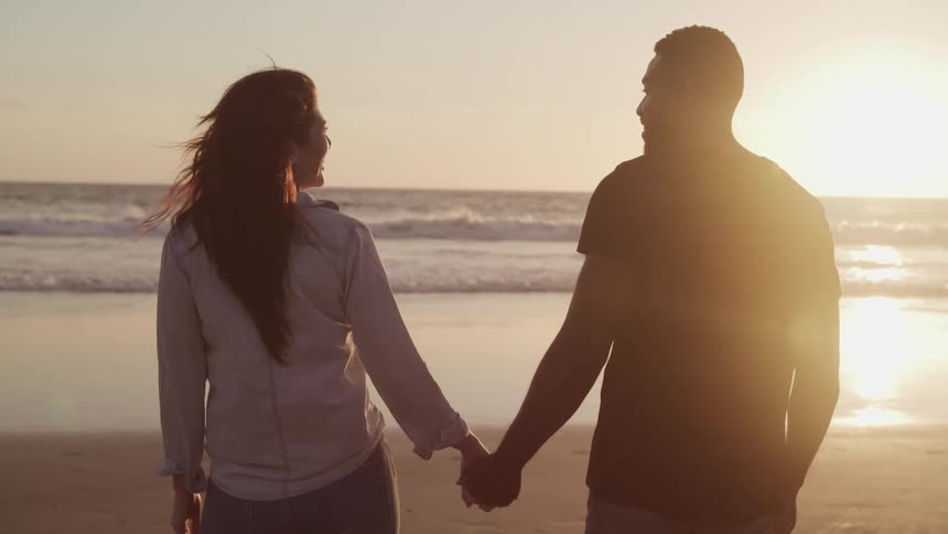 A man and woman smile at one another as they walk on a beach holding hands