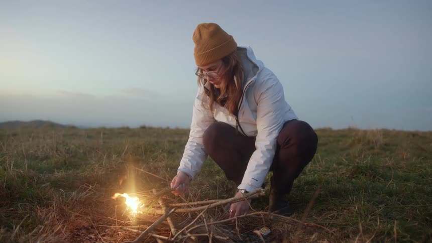 A woman is starting a fire outdoors in a grassland area