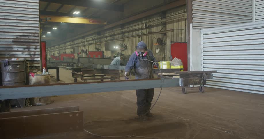 Worker wearing protective gear sprays beam near opening of factory as another worker welds