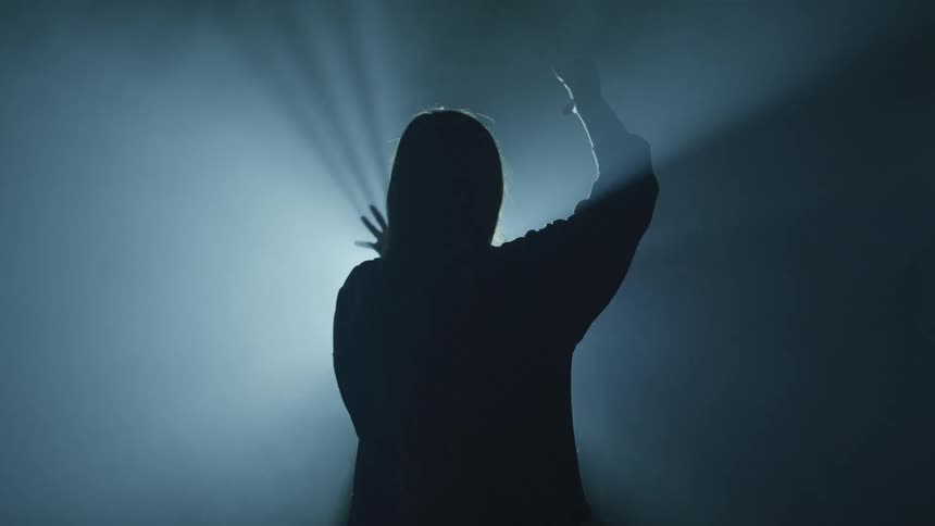 A woman stands in front of a flashing yellow light and waves her arms around in a dance move