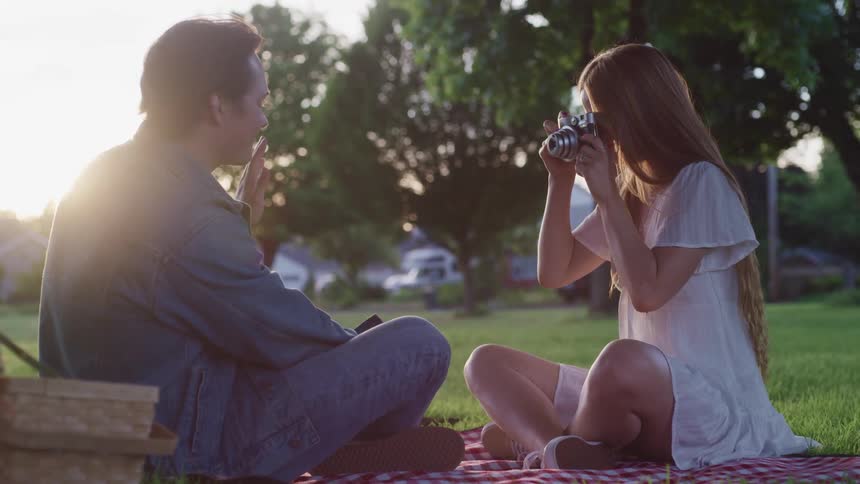 Young couple taking pictures of themselves with a vintage camera over the grass in a park at sunset