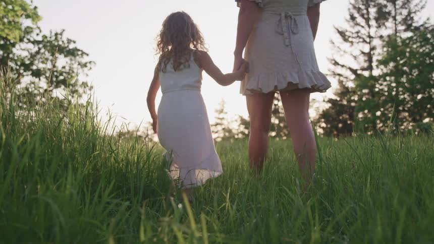 A woman and a girl walk through a field of grass while holding hands during the day