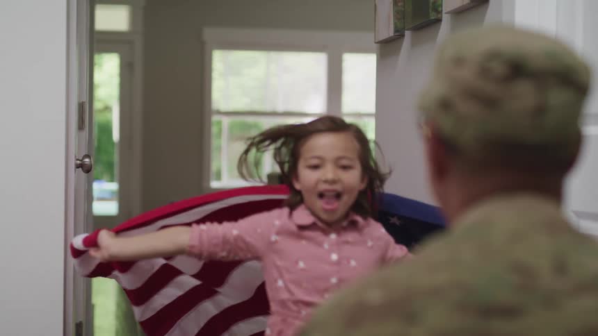 A young girl holding an american flag races to greet her father who is dressed in camouflage