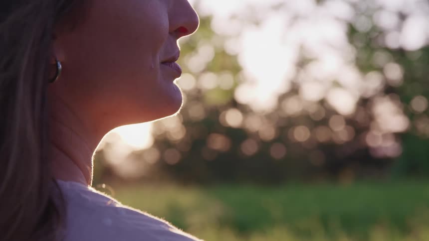 A woman stands outdoors with greenery and sunlight