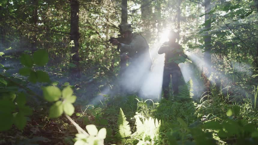Sunlight shines through tall trees in a forest as two military members stand with their guns drawn and pointed