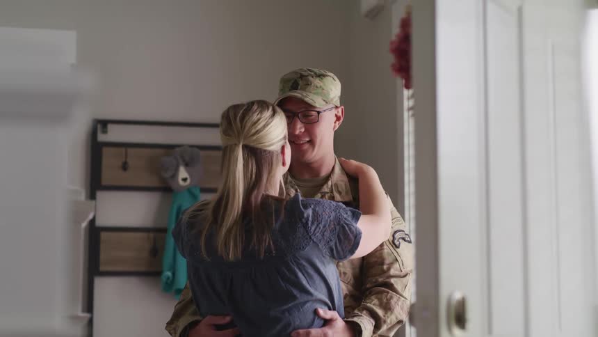 A male soldier embraces his wife upon arriving through the front door of a house