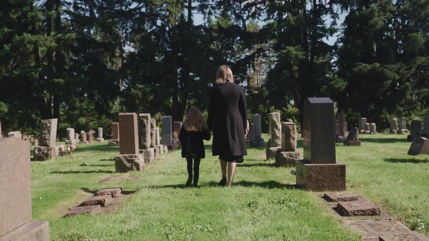 A woman and child holdings hands wearing black and walking in a cemetery