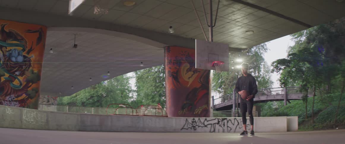 Young man playing basketball alone in a public court under a bridge in a park