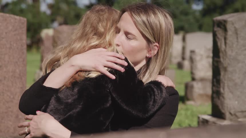 Mother and daughter hug each other in a cemetery in front of a grave