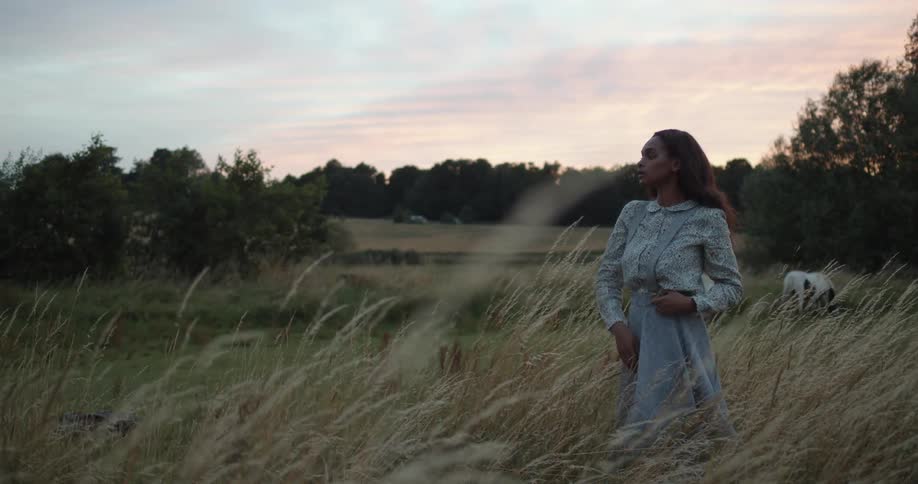 A woman standing out in tall grass in a meadow with horses in the distance runs her hand through her hair at sunset